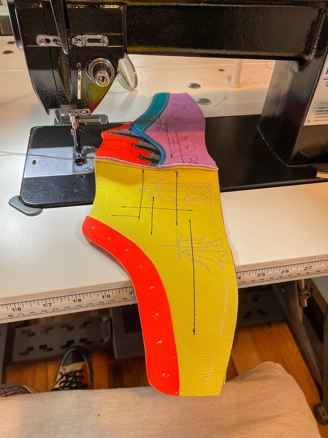 On a sewing machine,
the yellow high-top is sewn at the heal
to a lavender low-top and orange addition.
The yellow side has stitching of two moths,
and some straight black lines across,
with orange lacing panels.
The lavender side has a blue trim
and then silver-and-black stripes protruding
up and forward into the orange addition.