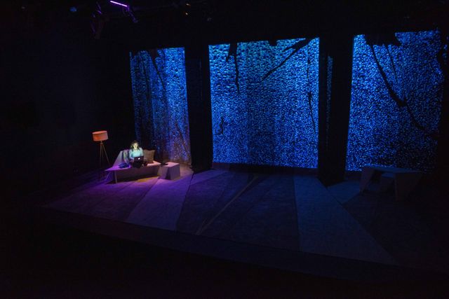 High side angle of a mostly bare stage, a woman is on a couch in a pool of light and the glow of her laptop -- large screens with rain projected behind her